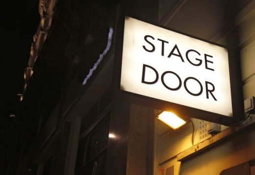 Valaistu kyltti, jossa lukee "Stage Door", kiinnitetään rakennuksen ulkopuolella olevaan seinään yöllä ja osoittaa sisäänkäynnin, jota tyypillisesti käyttävät esiintyjät tai henkilökunta.
