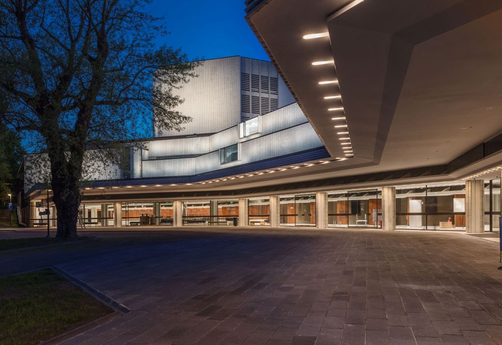 Moderni rakennus, jossa on suuret ikkunat ja geometrisia muotoja, valaistu hämärässä. Vasemmalla on puu, ja leveä, kivetty sisäänkäynti on valaistu kattovalaisimilla, mikä luo viihtyisän ja nykyaikaisen tunnelman.