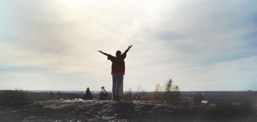 Henkilö seisoo kallion päällä kädet taivasta kohti kohotettuna ja katsoo pilviseen maisemaan, jossa on puita ja rakennuksia kaukana.