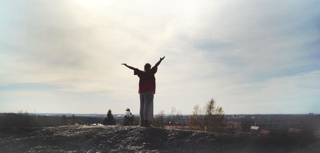 Henkilö seisoo kallioisella kukkulalla kädet taivasta kohti kohotettuna, siluetti pilvistä, kirkasta horisonttia vasten, ja kaukaisuudessa näkyy puita ja kattoja.