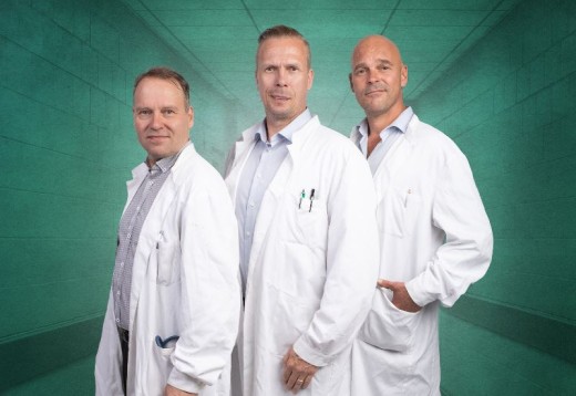 Kolme valkoisiin laboratoriotakkeihin pukeutunutta miestä seisoo vierekkäin käytävällä, jonka seinät ovat vihreät, ja katsoo luottavaisesti kameraan. Kahdella heistä on rintataskussaan kyniä ja työkaluja.