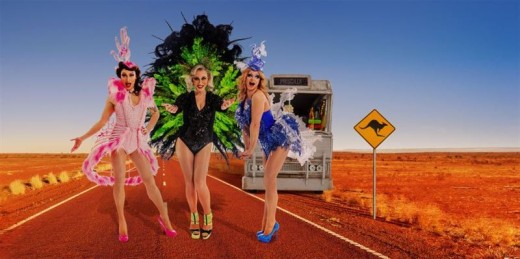 Kolme värikkäisiin, näyttäviin asuihin pukeutunutta ihmistä seisoo tyhjällä tiellä Australian takamaastossa. Näkyvissä on Priscilla-niminen bussi ja kengurun ylitysmerkki, taustalla punainen aavikko ja kirkkaansininen taivas.