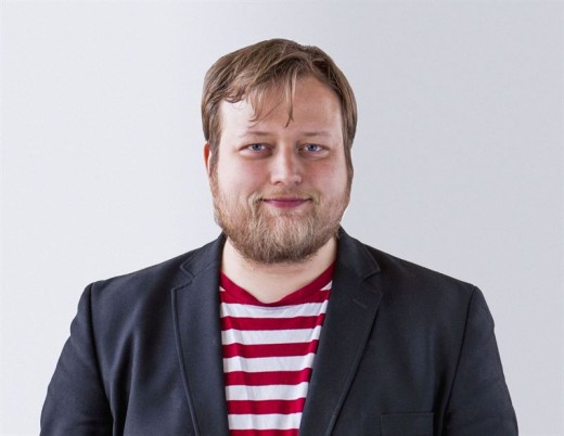 Vaaleanruskeat hiukset ja partainen mies seisoo valkoista taustaa vasten ja hymyilee hieman kameralle, yllään musta bleiseri ja punavalkoraidallinen paita.