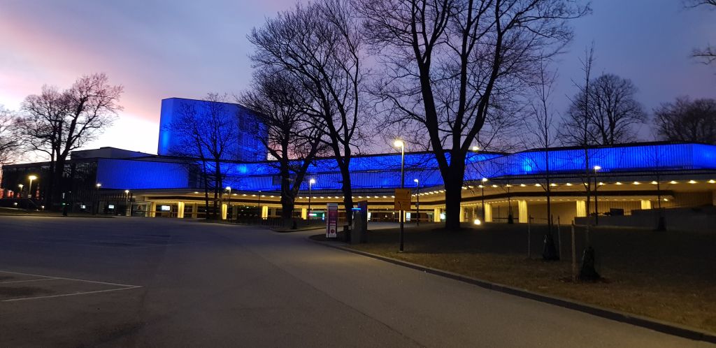 Moderni rakennus, joka on valaistu sinisillä valoilla iltahämärässä ja jota ympäröivät lehdettömät puut ja tyhjä parkkipaikka hämärän taivaan alla.