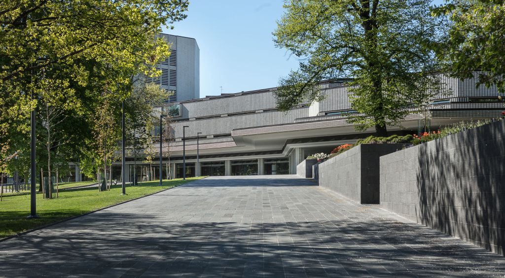 Leveä kivinen kävelytie johtaa moderniin, harmaaseen rakennukseen, jossa on suuret ikkunat ja jota ympäröivät vihreät puut ja maisemoitu ruoho kirkkaan sinisen taivaan alla.