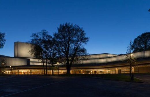 Moderni, matala rakennus, jonka lasiseinät on valaistu, on hämärässä puiden ympäröimä, etualalla on enimmäkseen tyhjä parkkipaikka ja yläpuolella on syvän sininen taivas.