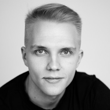 Nuori mies, jolla on lyhyet vaaleat hiukset ja vaaleat silmät, katsoo suoraan kameraan, ja hänellä on tumma paita vaaleaa taustaa vasten.