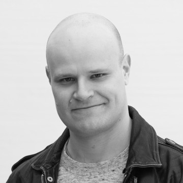 Mies, jolla on ajeltu pää ja hento hymy, pukeutuu vaaleaan villapaitaan ja tummaan takkiin. Tausta on tavallinen ja vaalea, ja kuva on mustavalkoinen.