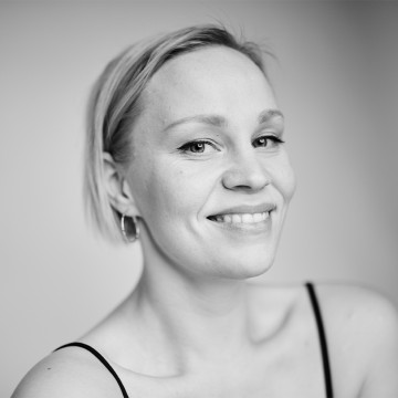 Mustavalkoinen muotokuva hymyilevästä naisesta, jolla on lyhyet vaaleat hiukset, korvakorut ja ohut nauhatoppi, ja joka katsoo hieman sivulle tavallista taustaa vasten.