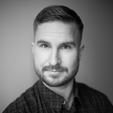 Mustavalkoinen muotokuva lyhythiuksisesta, parrakas mieshenkilöstä, jolla on ruudullinen nappipaita ja joka katsoo suoraan kameraan neutraali ilme.