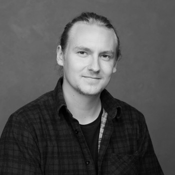 Vaaleaihoinen nuori mies, jolla on pitkät taakse sidotut hiukset, hymyilee pehmeästi. Hänellä on ruudullinen paita tumman t-paidan päällä ja hän istuu mustavalkoisessa muotokuvassa tavallista, neutraalia taustaa vasten.