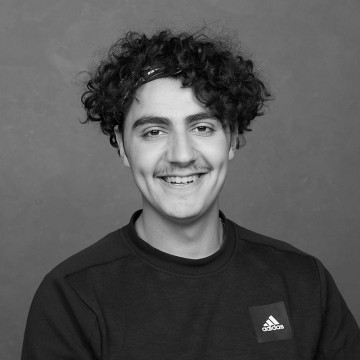 Nuori mies, jolla on kiharat hiukset ja viikset, tumma Adidas-verryttelypaita ja päähine, hymyilee kameralle harmaata taustaa vasten.