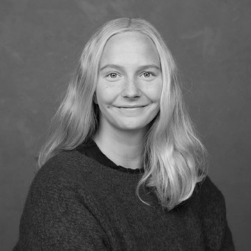 Mustavalkoinen muotokuva nuoresta henkilöstä, jolla on pitkät, vaaleat hiukset, yllään tumma villapaita, hymyilee pehmeästi ja poseeraa tavallisen taustan edessä.
