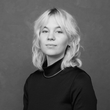 Nuori henkilö, jolla on vaaleat, aaltoilevat hiukset ja rauhallinen ilme, on pukeutunut mustaan poolopuseroon ja ohueen kaulakoruun, ja poseeraa mustavalkoisessa muotokuvassa tavallista, kuvioitua taustaa vasten.