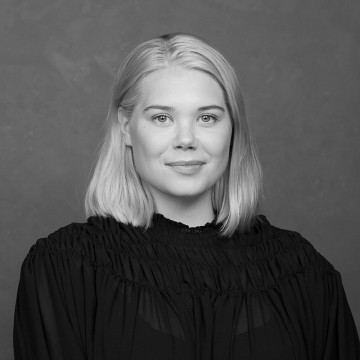 Mustavalkoinen muotokuva naisesta, jolla on suorat, hartioiden pituiset vaaleat hiukset, yllään tumma, kuvioitu toppi, katselee kameraan hymyillen tavallista taustaa vasten.