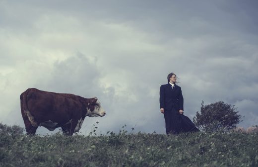 Pitkään mustaan takkiin pukeutunut henkilö seisoo ruohoisella kukkulalla pilvisen taivaan alla ja katsoo sivulle, kun taas ruskeavalkoinen lehmä seisoo lähellä ja katsoo myös samaan suuntaan.