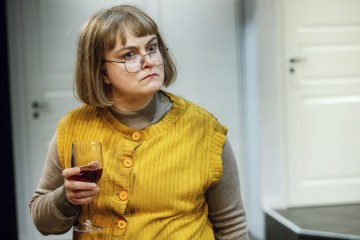 Lyhyet vaaleat hiukset ja silmälasit omaavalla naisella on keltainen liivi ja beige toppi, ja hänellä on kädessään punaviinilasi ja hän katsoo sivulle vakavalla ilmeellä. Taustalla näkyy valkoisia ovia.