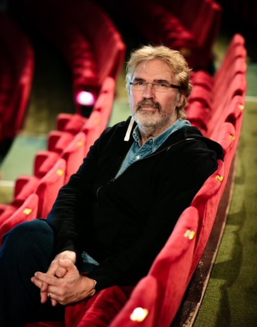 Silmälasipäinen herra istuu punaisella tuolilla teatterin katsomossa. Hän on ristinyt kätensä.