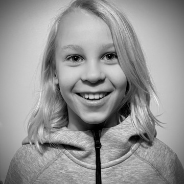 Mustavalkoinen valokuva hymyilevästä lapsesta, jolla on olkapäiden pituiset vaaleat hiukset, päällään vetoketjullinen huppari, kameraan päin, taustan ollessa tavallinen.