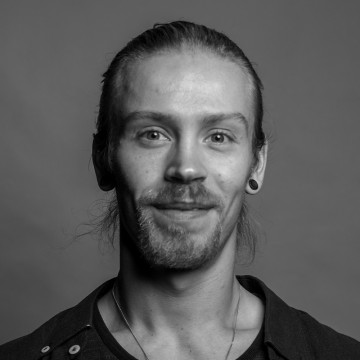 Mustavalkoinen muotokuva nuoresta miehestä, jolla on vaaleat kasvot, pitkät hiukset taaksepäin sidottuina, korvakorut ja tumma paita, hymyilee hieman kameralle. Tausta on yksinkertainen ja neutraali.
