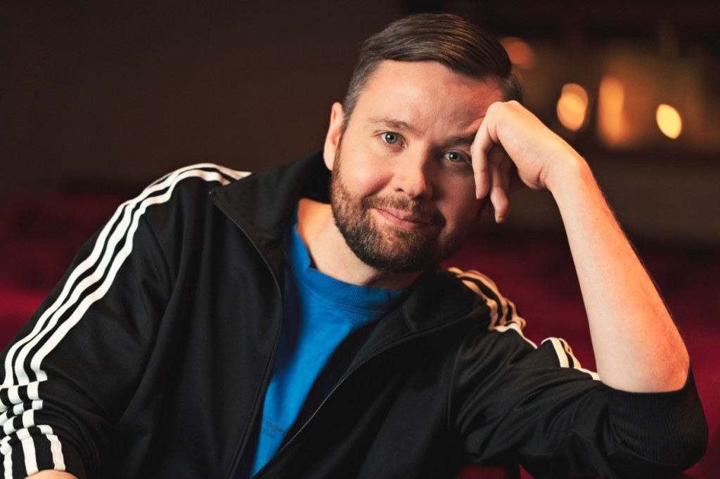 Parrakas mies nojaa vasempaan käteensä. Hänellä on verryttelypuvun takki ja sininen t-paita. Takana näkyy teatterin katsomo.