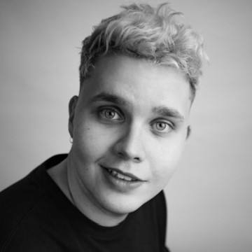 Henkilö, jolla on lyhyet, kiharat vaaleat hiukset ja vaaleat silmät, hymyilee kevyesti ja katsoo kameraan. Hänellä on yllään tumma toppi. Kuva on mustavalkoinen.