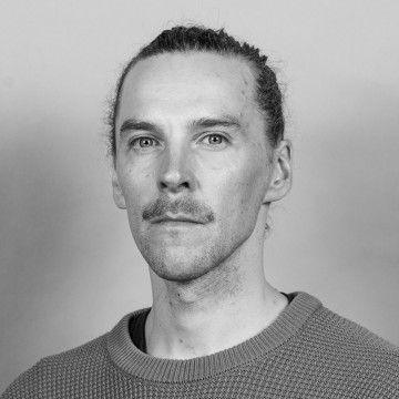 Mustavalkoinen muotokuva henkilöstä, jolla on vaalea iho, pitkät taakse vedetyt hiukset ja viikset, päällään neulepusero ja joka katsoo suoraan kameraan neutraali ilme.