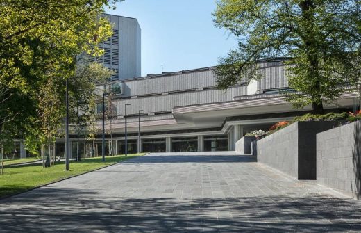 Moderni, harmaa kivirakennus, jossa on suuret ikkunat ja geometriset linjat, on aurinkoisena päivänä vihreiden puiden ja maisemoidun alueen ympäröimä.