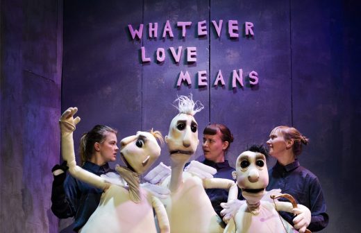 Neljä tummiin vaatteisiin pukeutunutta nukketeatteritaiteilijaa pitää suuria valkoisia nukkeja harmaan seinän edessä, jonka yläpuolella on vaaleanpunaisin kirjaimin teksti WHATEVER LOVE MEANS. Kohtaus on kirkkaasti valaistu ja teatraalinen.