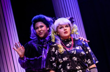 Kaksi värikkäisiin teatteripukuihin pukeutunutta henkilöä esiintyy lavalla. Toisella on tumma, röyhelöinen mekko ja violetit hiukset, kun taas toisella on vaaleat hiukset, joissa on vihreät korostukset, ja pandakuvioinen asu. He seisovat suurten, valkoisten pylväiden edessä.