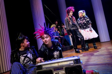 Ryhmä värikkäisiin punk-asuihin ja piikkikampauksiin pukeutuneita nuoria on vuorovaikutuksessa lavalla, kaksi istuu suuren boomboxin ääressä ja muut seisovat taustalla korkeiden, valkoisten pylväiden keskellä.