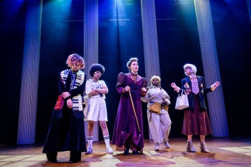 Viisi värikkäisiin, omalaatuisiin pukuihin pukeutunutta ihmistä seisoo lavalla, jossa on korkeat pylväät ja dramaattinen valaistus, ja näyttää esiintyvän teatteriesityksessä. Kukin henkilö poseeraa ainutlaatuisessa asennossa, mikä viittaa erilaisiin hahmoihin.
