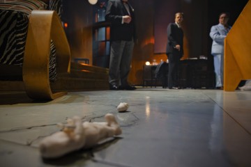 Etualalla lattialla makaa kaksi pientä rikkinäistä hahmoa, ja taustalla seisoo kolme muodollisesti pukeutunutta henkilöä keskustelemassa hämärässä, tyylikkäässä huoneessa, jossa on modernit huonekalut.