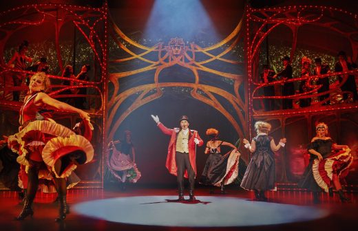Teatterillinen näyttämökohtaus, jossa punatakkinen sormusmestari on keskellä näyttämöä, ja häntä ympäröivät tanssijat värikkäissä can-can-tyylisissä puvuissa dramaattisessa valaistuksessa ja koristeellisessa, punakultaisessa lavastuksessa, jonka taustalla on parvekkeella esiintyviä esiintyjiä.