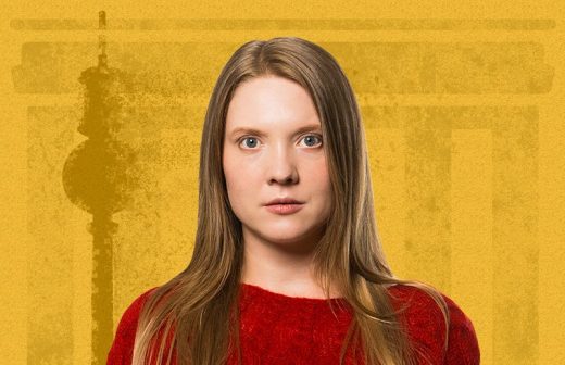 Nuori nainen, jolla on pitkät suorat hiukset ja punainen villapaita, seisoo keltaisella taustalla, jossa on arkkitehtonisia muotoja. Hän katsoo suoraan kameraan neutraalilla ilmeellä.