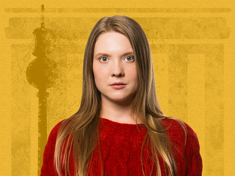 Nainen, jolla on pitkät suorat vaaleat hiukset ja punainen villapaita, tuijottaa suoraan kameraan. Taustalla on keltainen, haalistunut arkkitehtoninen kuvio.