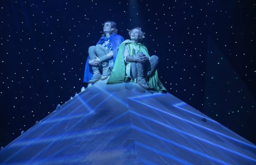 Kaksi sinisiin ja vihreisiin viittoihin pukeutunutta ihmistä istuu ristissä pyramidin muotoisen rakennelman päällä, jota ympäröi tähtitaivas ja dramaattinen sininen valaistus.