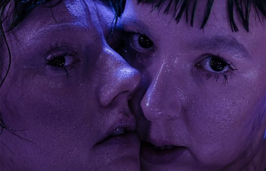 Kaksi ihmistä, joilla on märkä iho ja voimakkaat ilmeet, ovat tiiviisti kameraan päin violetissa valaistuksessa, heidän kasvonsa lähes koskettavat toisiaan ja heidän silmänsä ovat avoinna, mikä luo dramaattisen, intiimin tunnelman.
