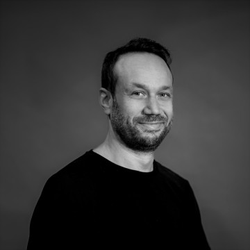 Mustavalkoinen kuva parrakkaasta miehestä, joka katsoo suoraan kameraan hymyillen hieman. Hänellä on musta paita.