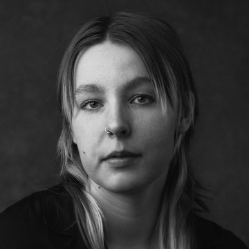 Mustavalkoinen muotokuva nuoresta henkilöstä, jolla on suorat hiukset, nenärengas ja neutraali ilme, ja joka katsoo suoraan kameraan tummaa, yksinkertaista taustaa vasten.