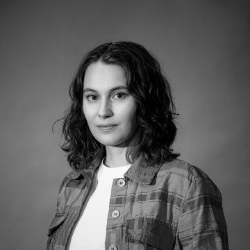 Mustavalkoinen muotokuva naisesta, jolla on hartioiden pituiset aaltoilevat hiukset, yllään ruudullinen paita ja tavallinen toppi, ja joka katsoo rauhallisesti kameraan tavallista taustaa vasten.