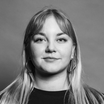 Mustavalkoinen muotokuva nuoresta naisesta, jolla on pitkät, suorat hiukset, korvakorut ja tumma toppi, ja joka katsoo suoraan kameraan neutraali ilme taustaa vasten.