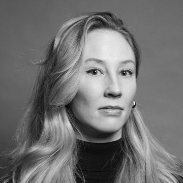 Mustavalkoinen muotokuva naisesta, jolla on pitkät, vaaleat, aaltoilevat hiukset, yllään tumma poolopaita ja pienet korvakorut, katselee suoraan kameraan neutraali ilme.