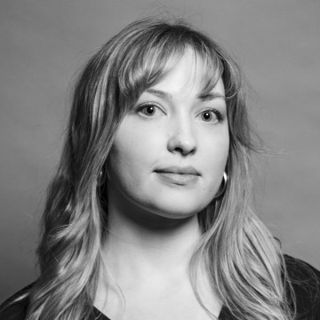 Mustavalkoinen muotokuva nuoresta naisesta, jolla on pitkät, aaltoilevat hiukset, korvakorut ja tumma toppi, katselee hieman kameran ohi neutraali ilme tavallista taustaa vasten.