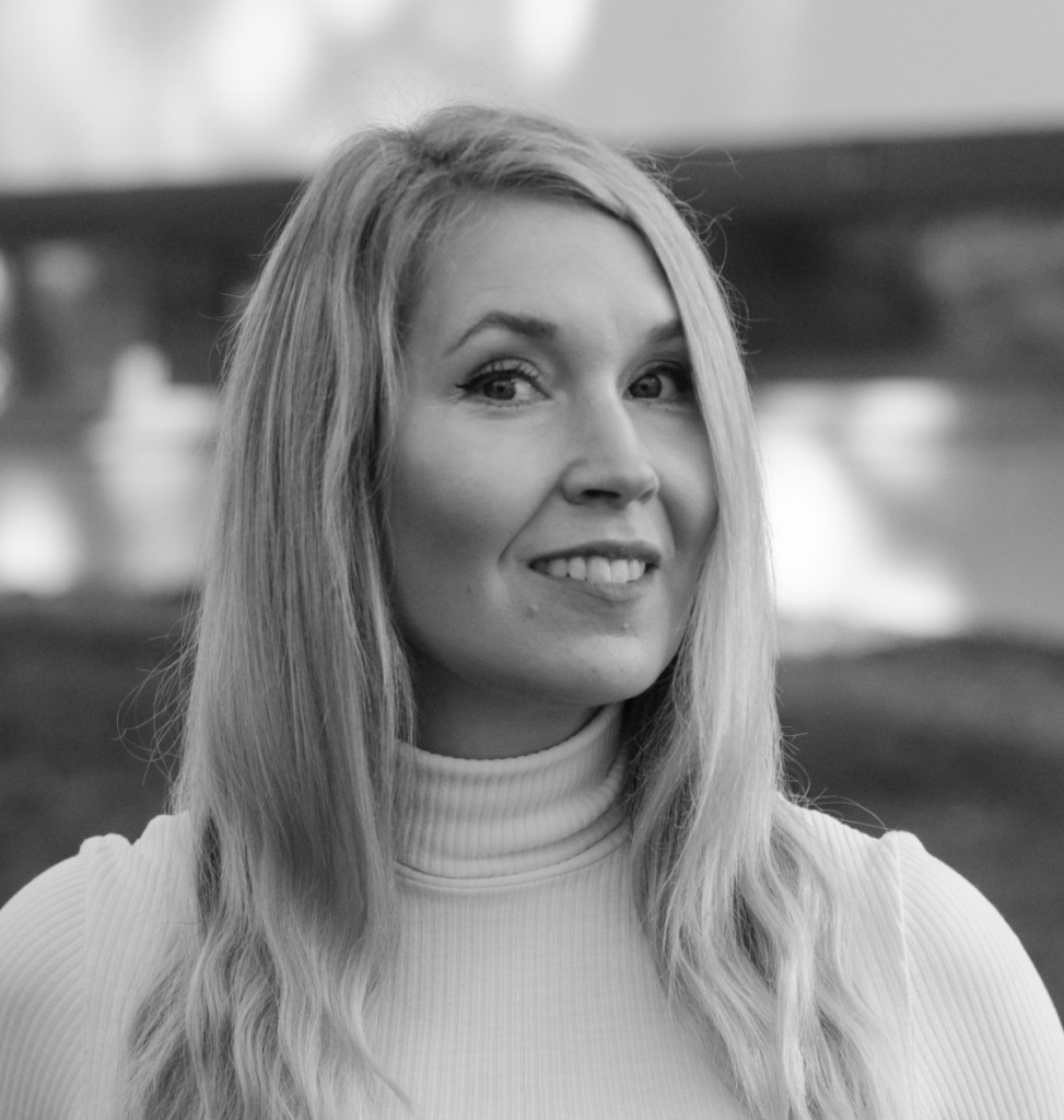 Mustavalkoinen muotokuva naisesta, jolla on pitkät, vaaleat hiukset ja vaalea poolopaita, hymyilee hieman ja katsoo kameraan päin, taustalla on sumea ulkoilma.