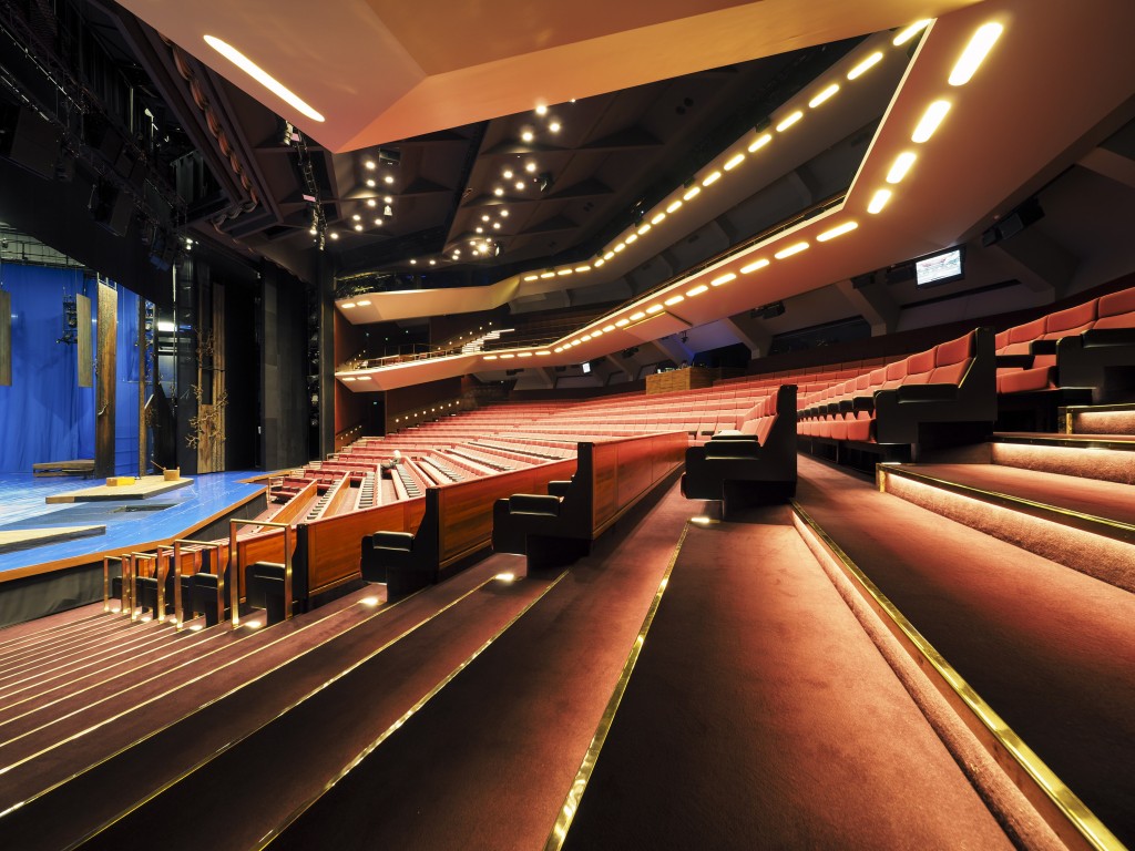 Nykyaikaisen teatterin sisätila, jossa on punaiset pehmustetut istuimet, valaistut portaat ja näkymä näyttämölle vasemmalla. Lämmin valaistus korostaa tyylikästä, nykyaikaista arkkitehtuuria ja tilavaa istumajärjestystä.