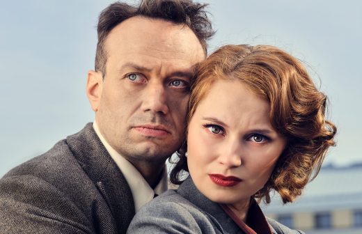 Mies ja nainen, jotka ovat pukeutuneet vintage-tyylisiin vaatteisiin, tuijottavat intensiivisesti kameraan ja seisovat lähekkäin ulkona sumealla taustalla.