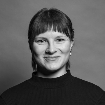 Mustavalkoinen muotokuva henkilöstä, jolla on lyhyet, suorat hiukset ja otsatukka, yllään tumma poolopusero, pienet korvakorut ja nenärengas, hymyilee pehmeästi kameralle tavallista taustaa vasten.