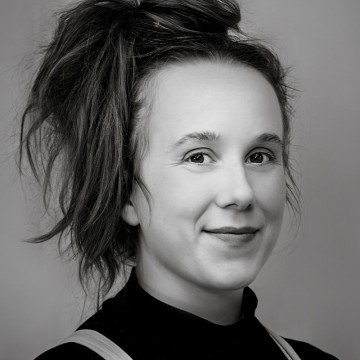 Mustavalkoinen muotokuva nuoresta naisesta, jolla on pitkät hiukset sidottuna korkeaan sotkuiseen pussiin, päällään tumma poolopaita ja joka hymyilee lempeästi kameralle.