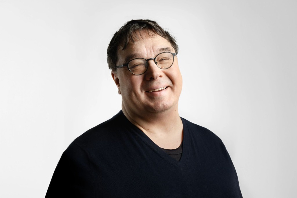 Keski-ikäinen henkilö, jolla on lyhyet tummat hiukset ja silmälasit, hymyilee lempeästi, kun hänellä on yllään tumma V-kaula-aukkoinen paita vaaleanharmaata taustaa vasten.
